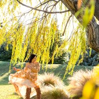 A brunette woman wears a floral dress which she is moving the skirt of, she stands beneath a willow tree and the frame is filled with golden light