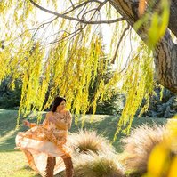 A brunette woman wears a floral dress which she is moving the skirt of, she stands beneath a willow tree and the frame is filled with golden light