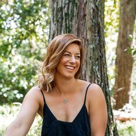 A slim yoga instructor sits under a tree on a sunny day, she is laughing at something off camera