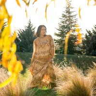 A brunette woman is framed by the branches of a willow tree, she wears a floral dress
