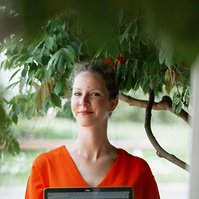 A woman in an orange dress stands under a tree as she proudly displays her laptop screen to the photographer