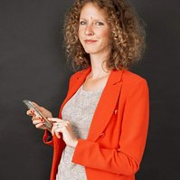 A curly haired woman smiles knowingly at the camera, her finger paused mid scroll on her phone, a reflection of her face visible on a table