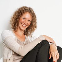 A slim woman with red curly hair sits on a white chair and smiles directly into the camera