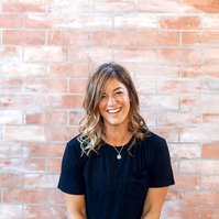 A female yoga teacher dressed all in black sits on a bolster in front of a brick wall