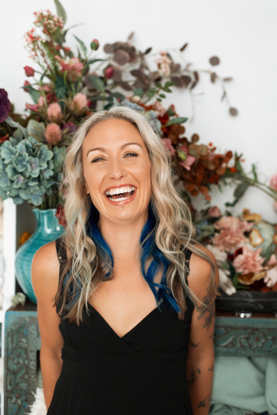 A woman with blue and blonde hair laughs, a large floral arrangement behind her