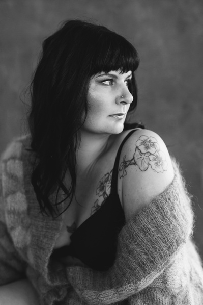 A sultry brunette sits wrapped in a jumper, looking over her tattooed shoulder