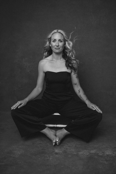 A quirky blonde woman rests on a stool, toes touching with her hands resting on wide knees as if she is in a yoga asana