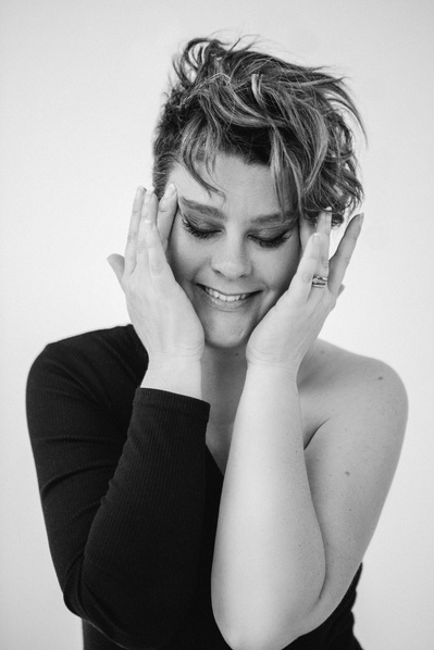A woman with short brown hair and an asymmetrical bodysuit touches her temples and smiles downward