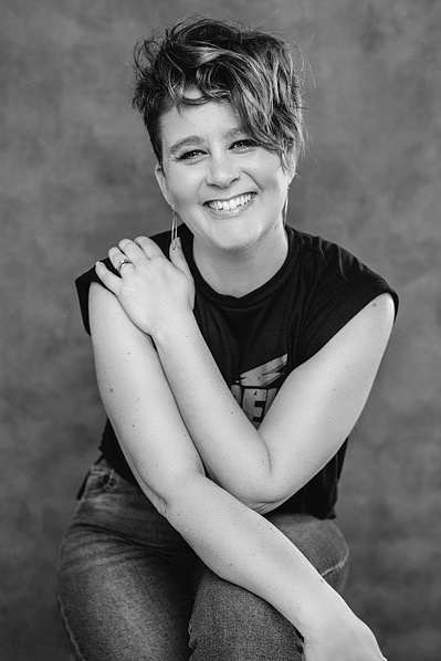 A woman smiles at the camera, short hair falling in her eyes. She wears jeans and a funky t-shirt