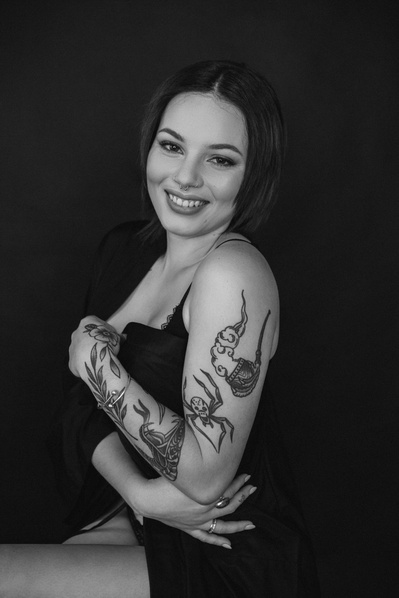 A young tattooed brunette wraps a dark sheet around herself and smiles at the camera