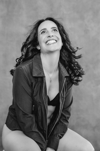 A slim woman with long brown hair wears leather jacket over a bra, laughing