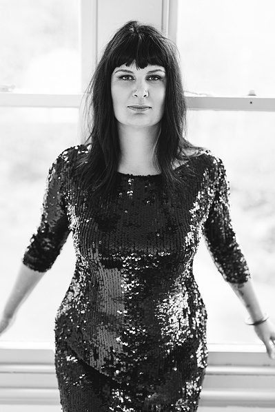 A striking brunette with a funky fringe leans back on a windowsill, wearing a sequinned dress