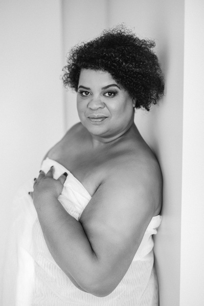 A curvaceous black woman leans against a wall wrapped in a white sheet giving the camera a sultry look