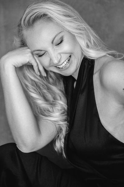 A close up of a blonde woman, resting her cheek on her hand as she smiles with her eyes closed