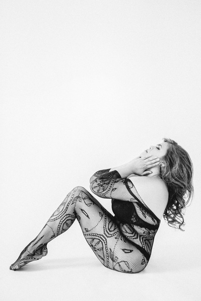A woman wears a lace bodysuit, sitting with her back arched and knees up