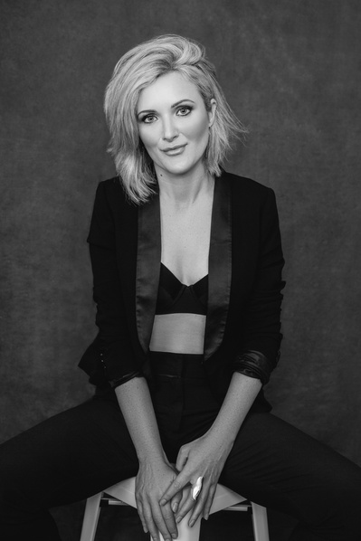 Striking blonde woman slouches on a stool wearing a bra and a black blazer
