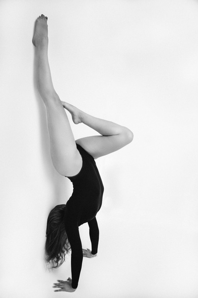 A slim woman wearing a bodysuit does a handstand against a white wall, one leg bent with toe pointed