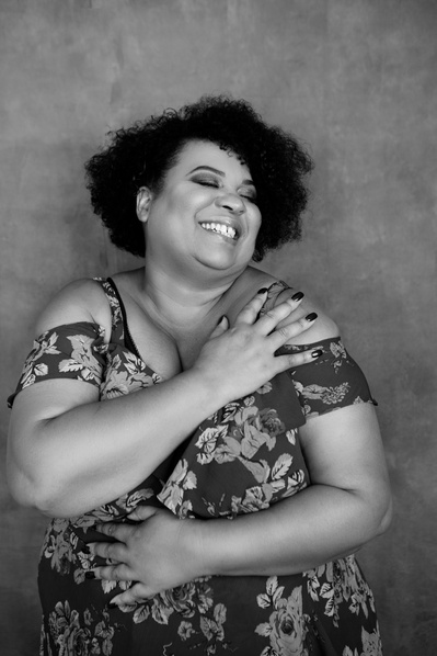 A curvaceous woman wearing a floral dress embraces herself with closed eyes, a smile on her face