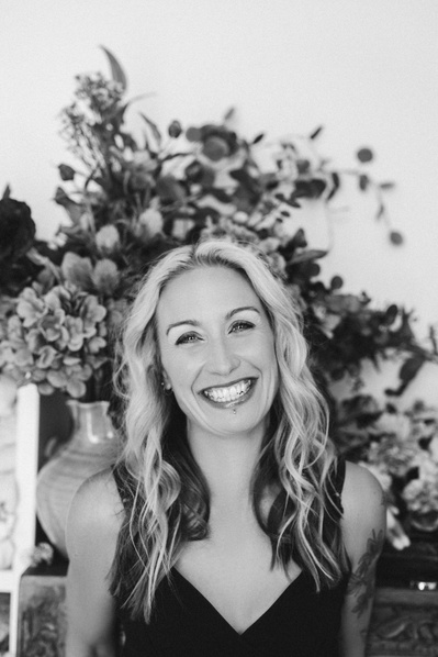 A blonde woman smiles at the camera, behind her is a large floral display