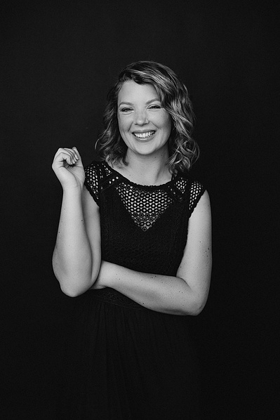 A glamourous young woman with curls smiles at the camera with one arm crossed over her torso