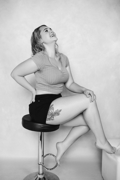 A young woman sits on a black stool, back arched as she laughs broadly