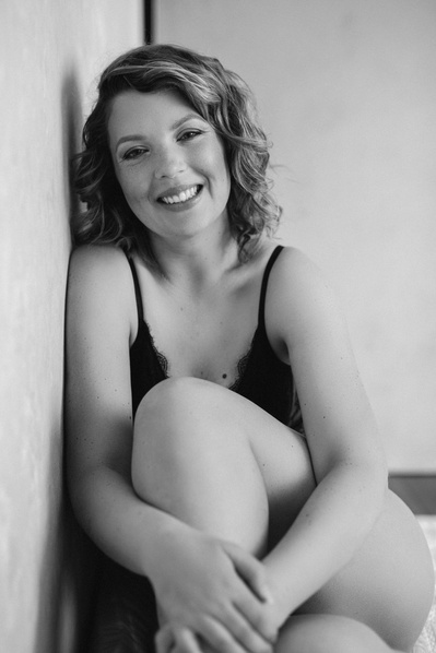 A woman with curly hair sits down, leans against a wall and is smiling