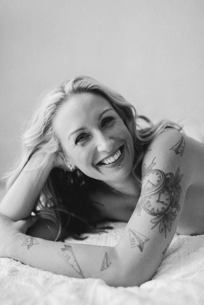 A blond woman smiles as she lays on a bed, her arms tattoos visible