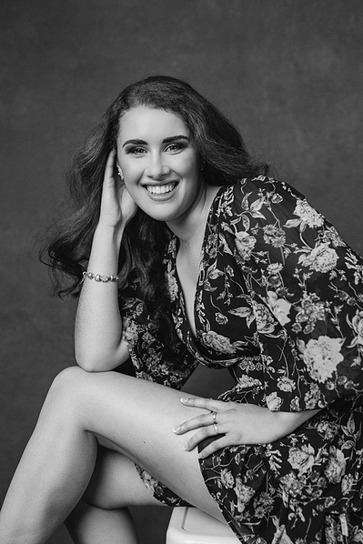 A young woman with long dark hair poses in a floral dress, hand supporting her head as she smiles