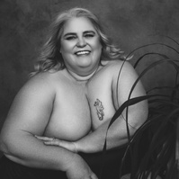 A curvaceous woman poses next to a large houseplant, she is smiling