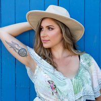A young woman leans against a blue door, an arm behind her head displaying her tattoo