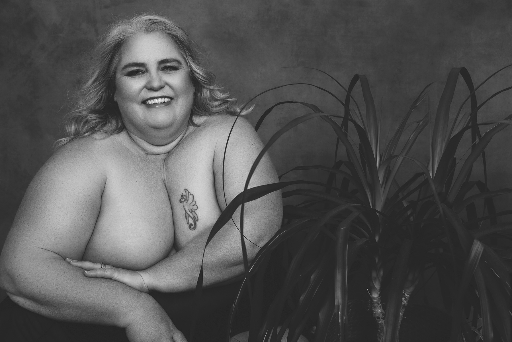 A curvaceous woman poses next to a large houseplant, she is smiling
