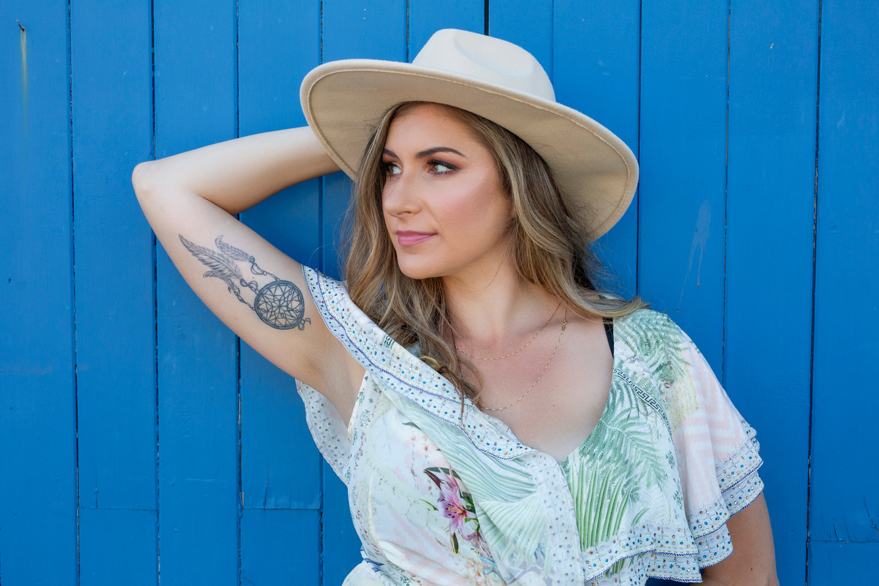 A young woman leans against a blue door, an arm behind her head displaying her tattoo
