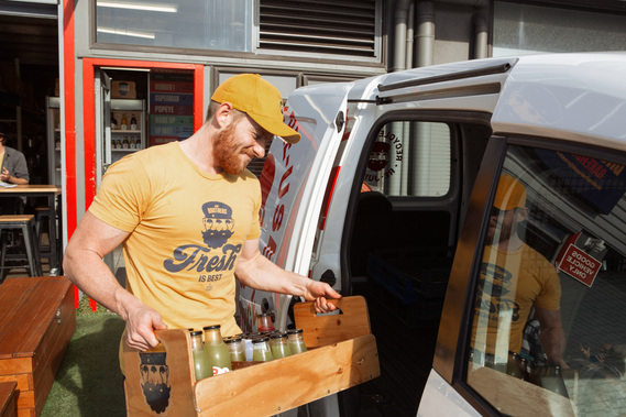 A worker loads a crate of smoothies into a van, he wears a yellow tshirt and cap and his muscles are visible