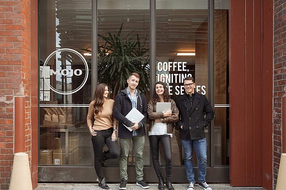 Four colleagues stand against the glass windows of their office, they are laughing together