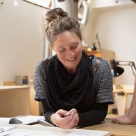 A woman with brown hair smiles during a meeting, her male co-worker leans on the table with only his torso in shot