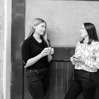 Two female colleagues stand with coffee cups in a hallway discussing their work
