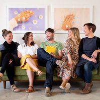 Five colleagues sit on a green sofa in their workspace, they look relaxed and are smiling at one another