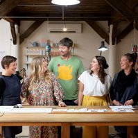 A team of five people smile at one another. They are all behind a wooden desk located in a jewellery workshop