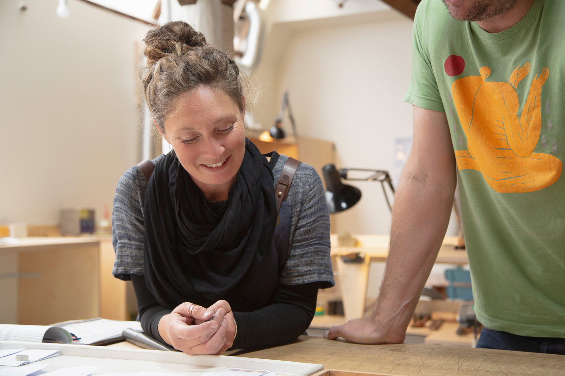 A woman with brown hair smiles during a meeting, her male co-worker leans on the table with only his torso in shot