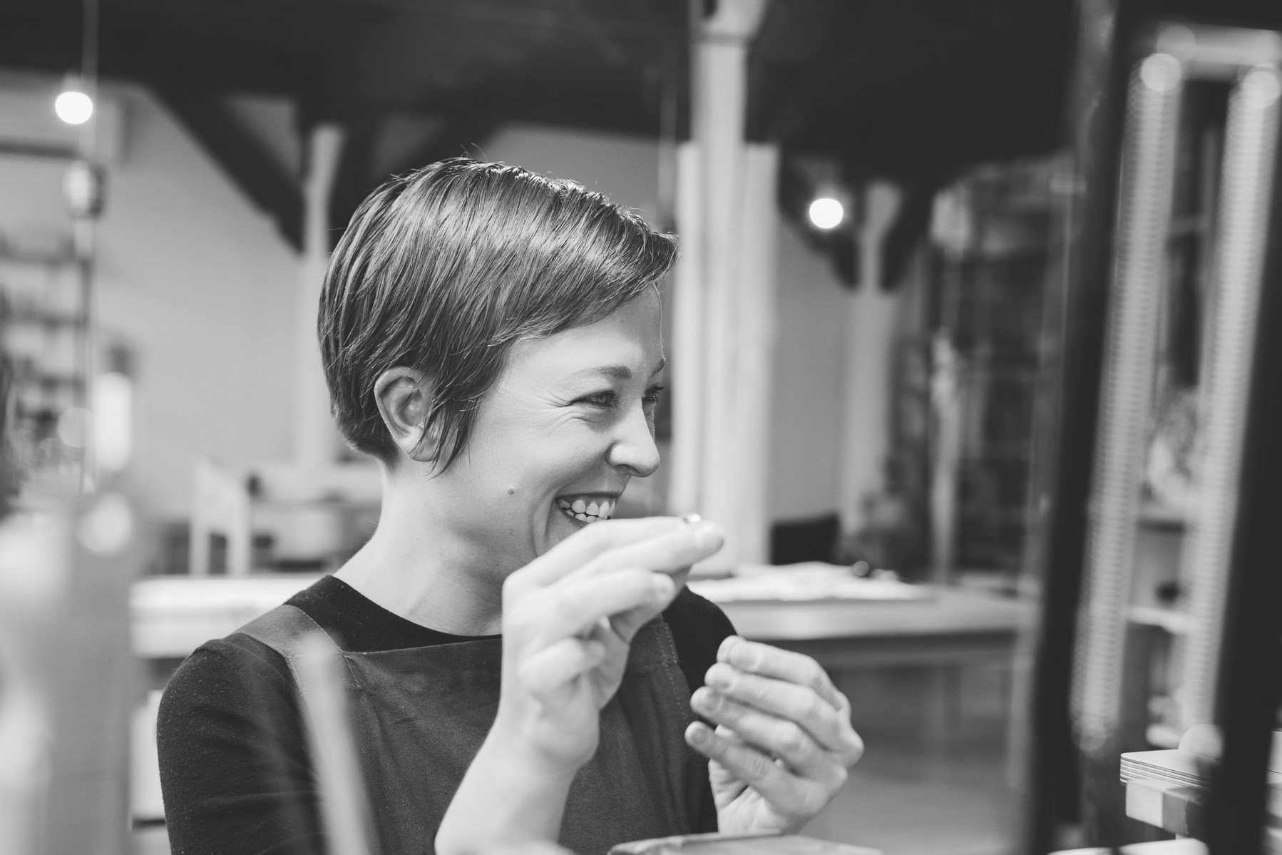 A woman with short brown hair smiles at something off camera as she works on designing a handmade ring