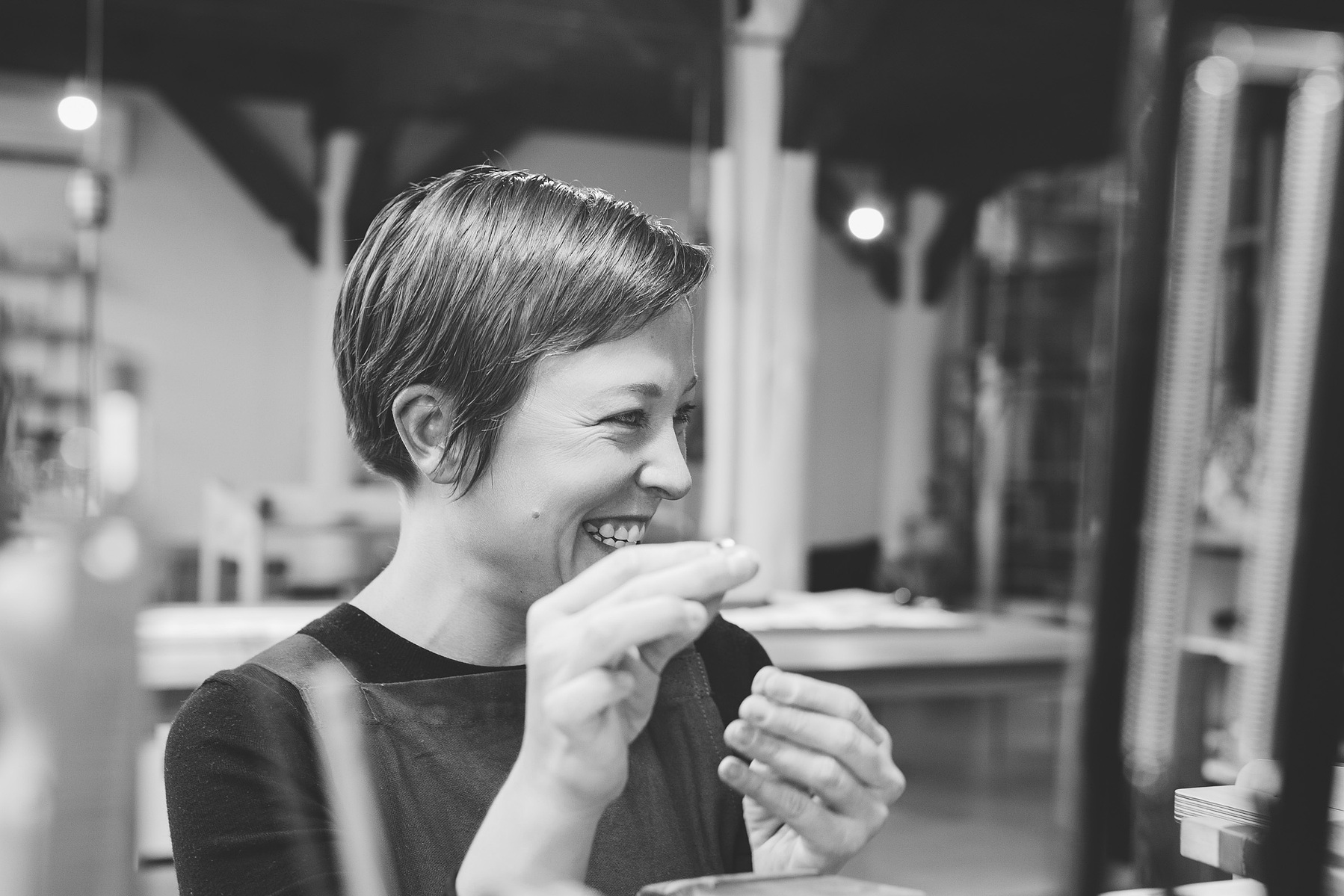 A woman with short brown hair smiles at something off camera as she works on designing a handmade ring