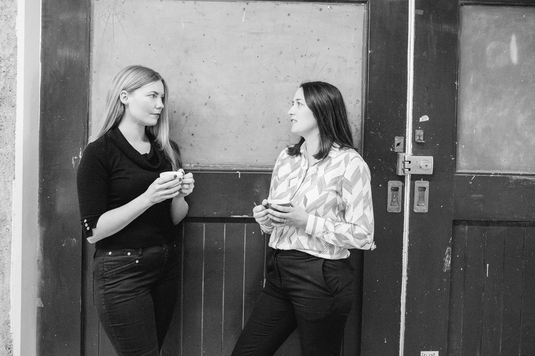 Two female colleagues stand with coffee cups in a hallway discussing their work
