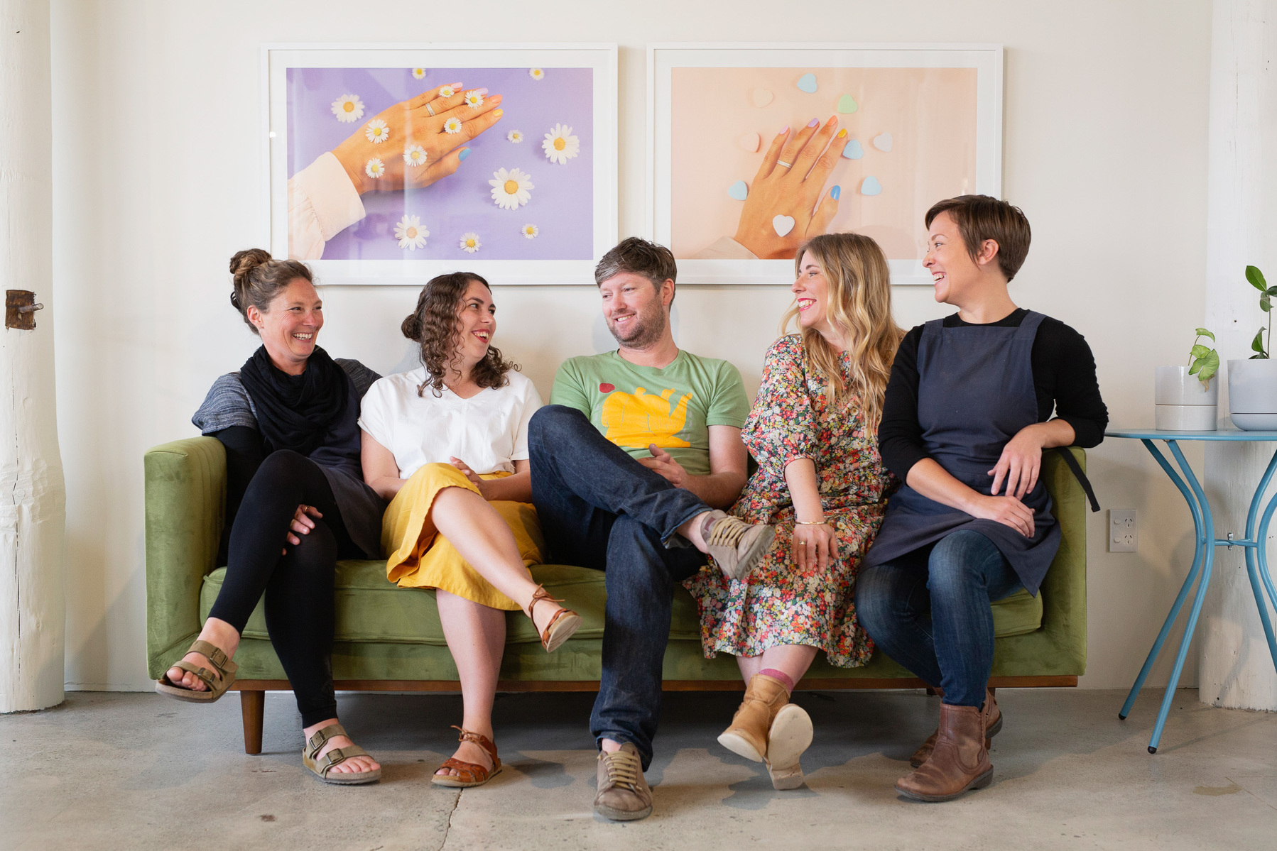 Five colleagues sit on a green sofa in their workspace, they look relaxed and are smiling at one another