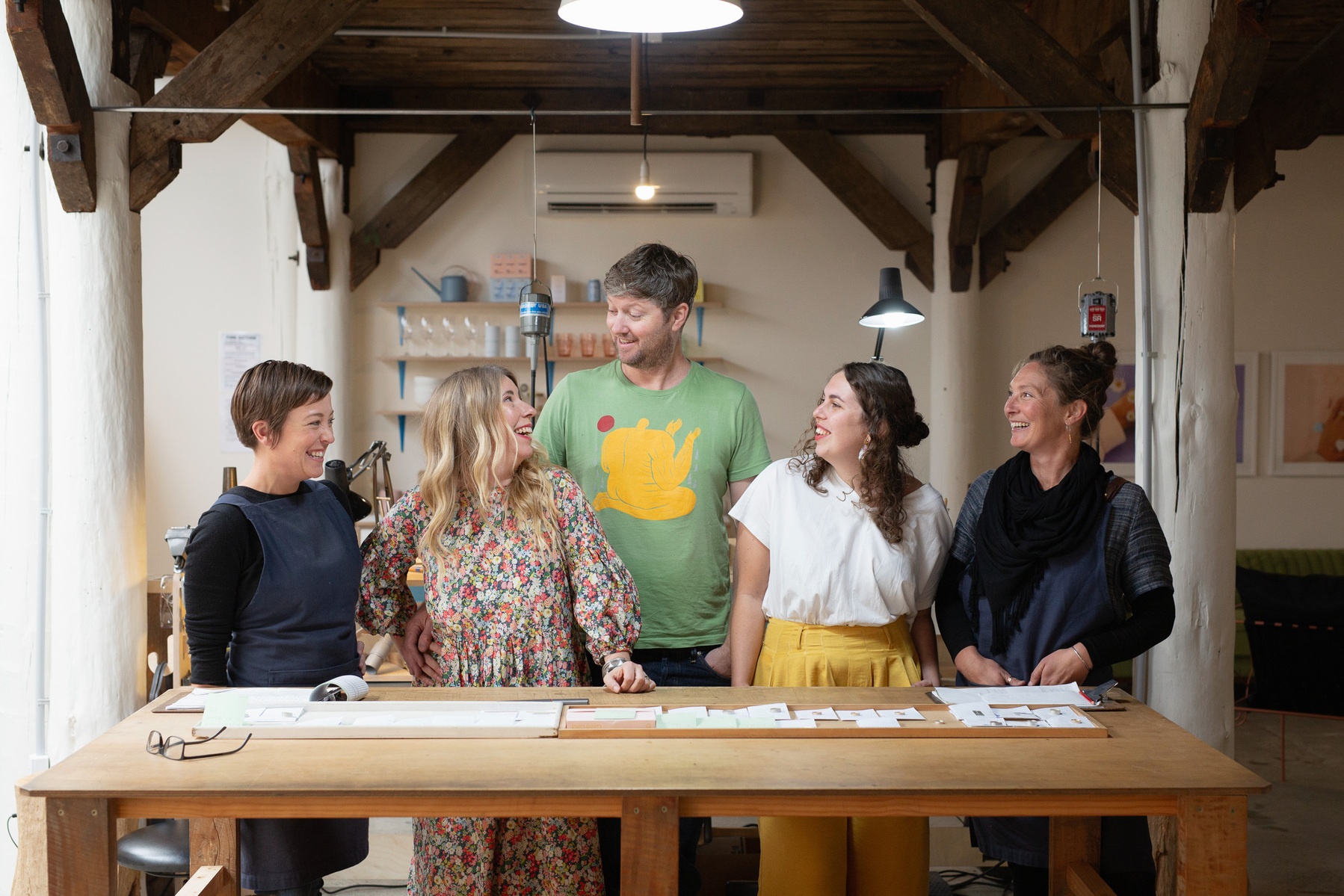A team of five people smile at one another. They are all behind a wooden desk located in a jewellery workshop