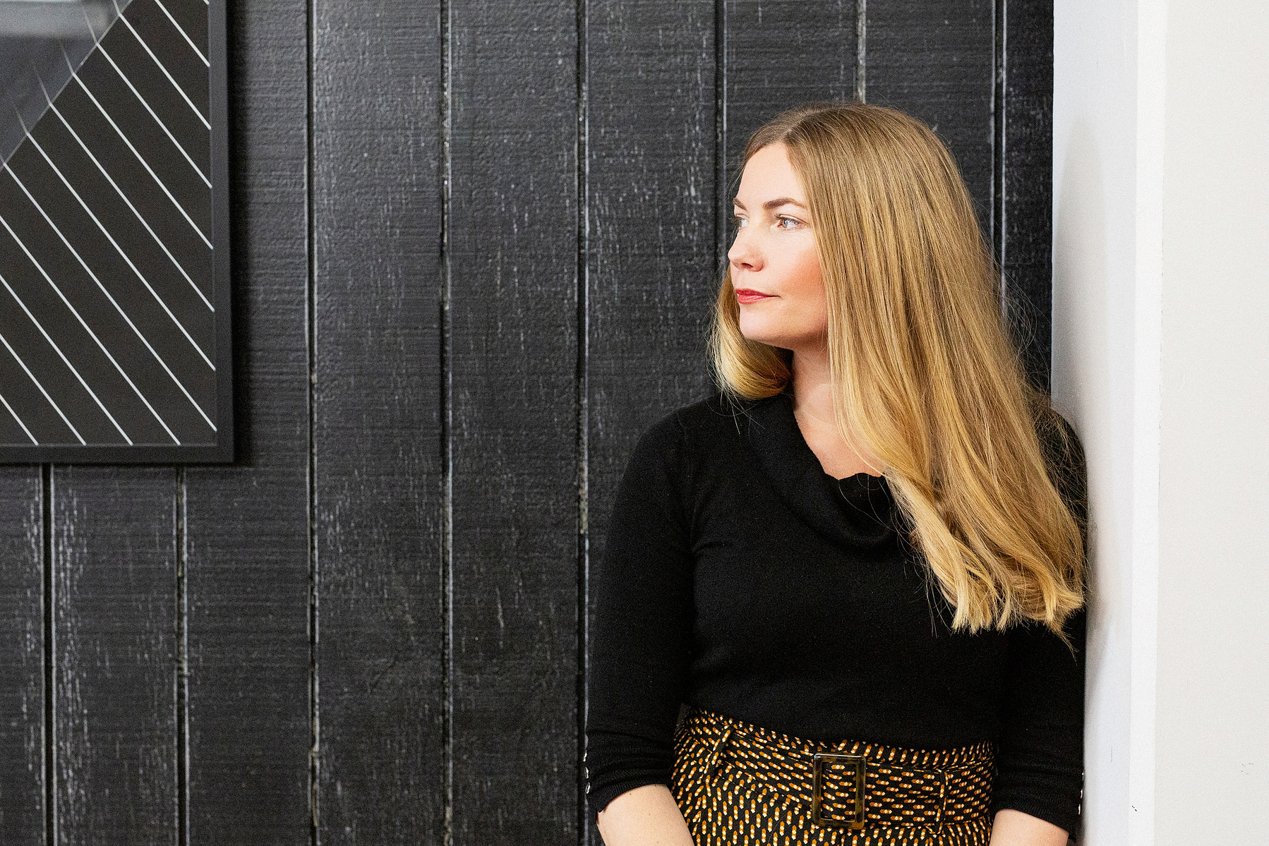 A blonde woman leans against a doorway and looks off camera to her right