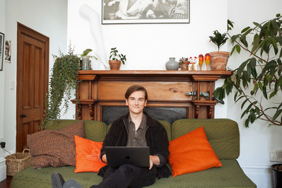 A young brunette man sits in a living room on a green sofa with a laptop on his lap
