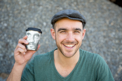 A young man wearing a flat cap holds up a cup of takeaway coffee and smiles