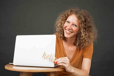 A curly haired smiling woman sits at a desk with her laptop open. She holds a cut out of letter spelling 'hello'