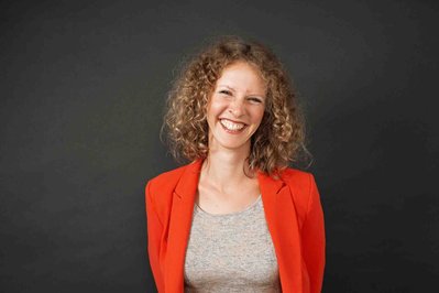 A woman with curly hair and an orange jacket smiles widely into the camera