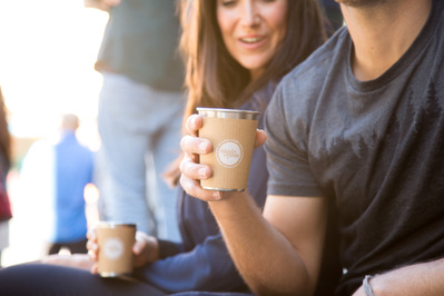 Two people enjoy takeaway coffee outside, a busy background full of people is visible